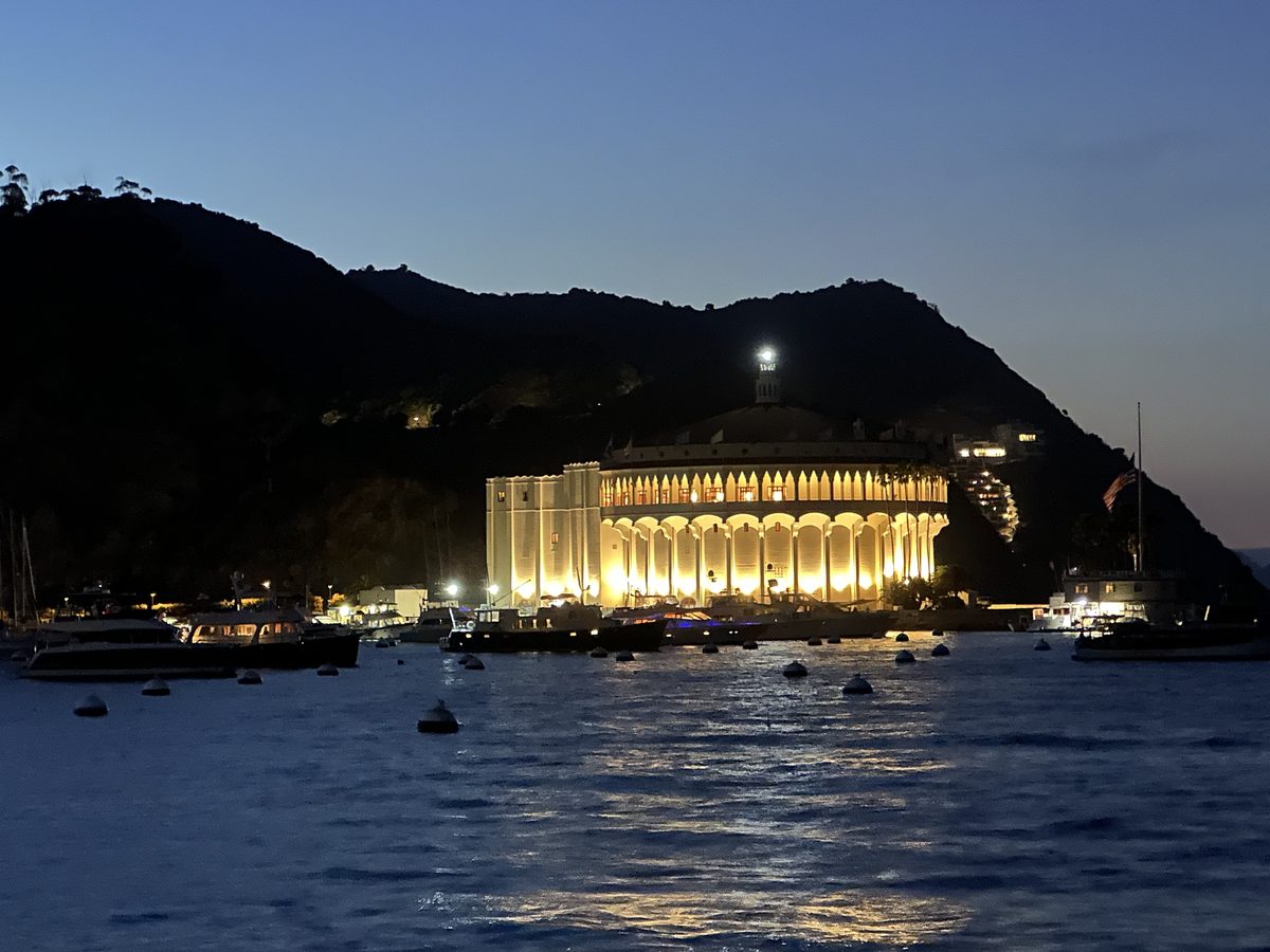 The Avalon Casino at blue hour — the building glows warm gold against a deep blue twilight sky, boats in the harbor below
