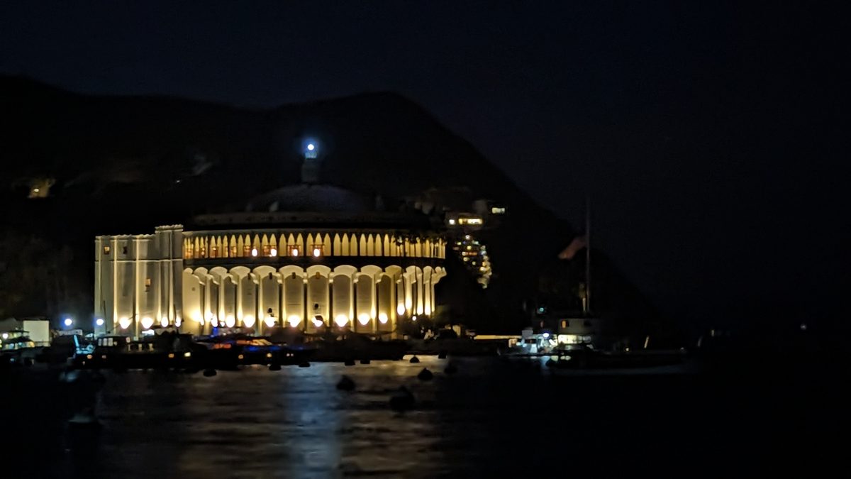 The Avalon Casino illuminated at night — its white colonnade glowing against the dark hillside, reflected in the harbor water