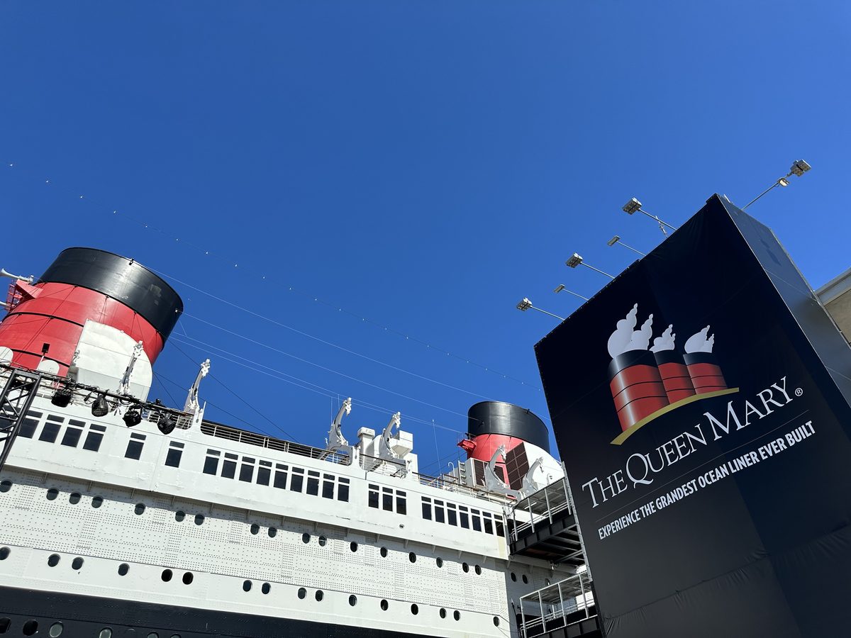 Looking up at the Queen Mary from the dock — two red-and-black funnels against a deep blue sky, with the Queen Mary sign in the foreground