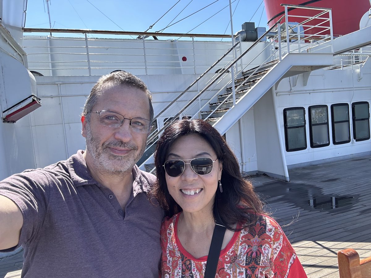 Eddy and Ako selfie on the Queen Mary's sun deck — the red funnel behind them, clear blue sky