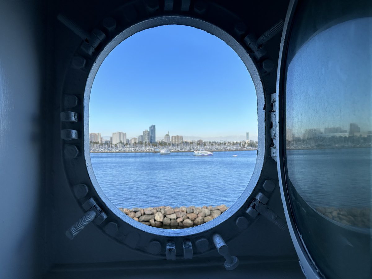 Two steel portholes side by side on the Queen Mary, one looking out to Long Beach harbor and skyline