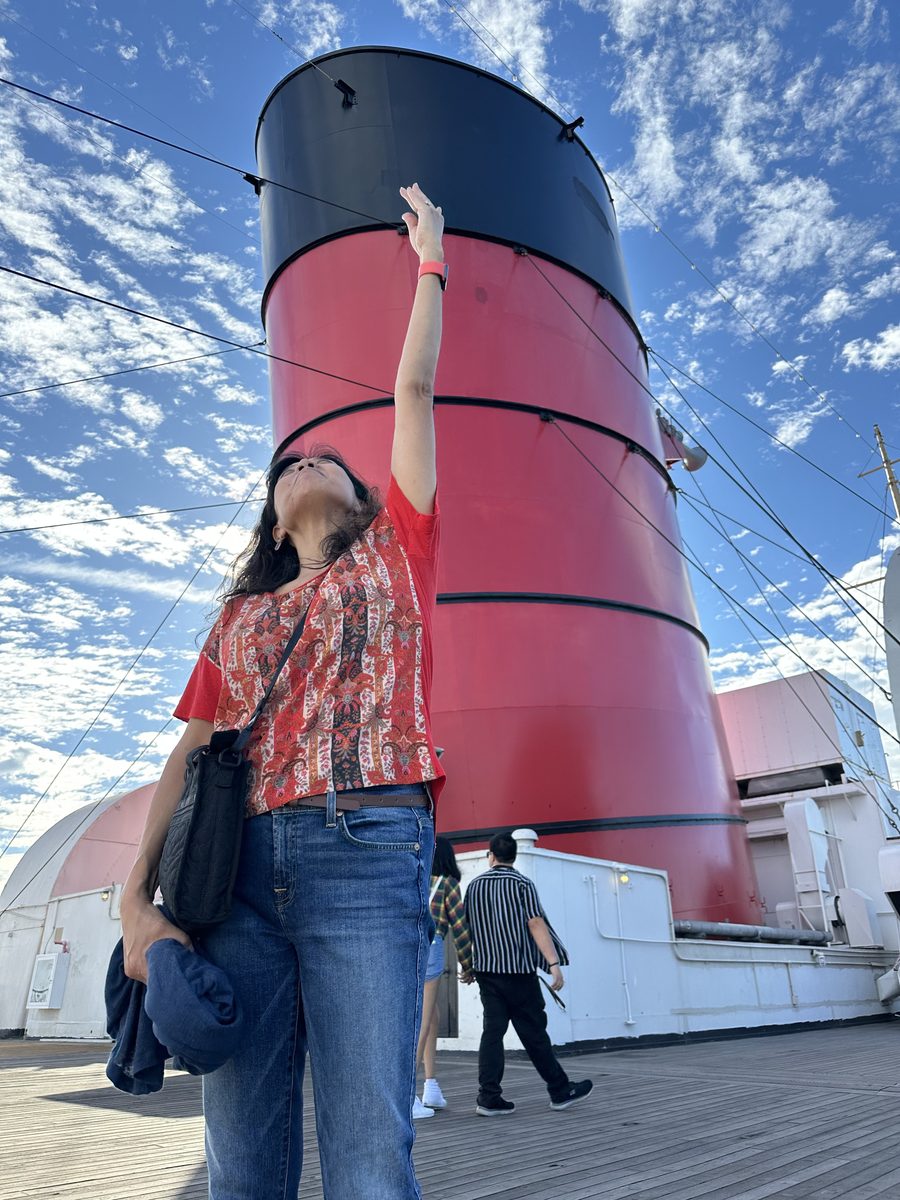 Ako reaching up toward the giant red Queen Mary funnel with one hand, looking up at it against a blue sky