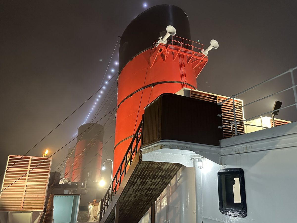 The Queen Mary's red funnel glowing at night through sea fog, deck equipment and a second funnel visible behind