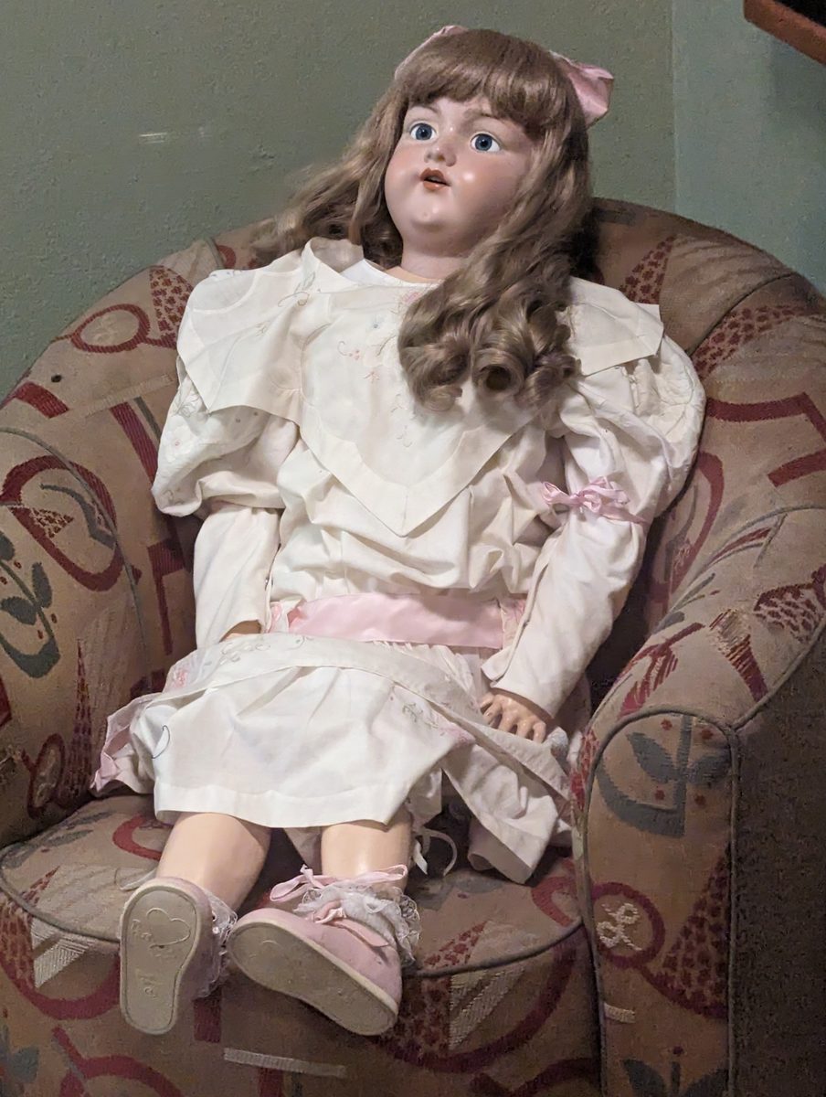 A large antique porcelain doll in a Victorian dress, seated in a chair on the Queen Mary