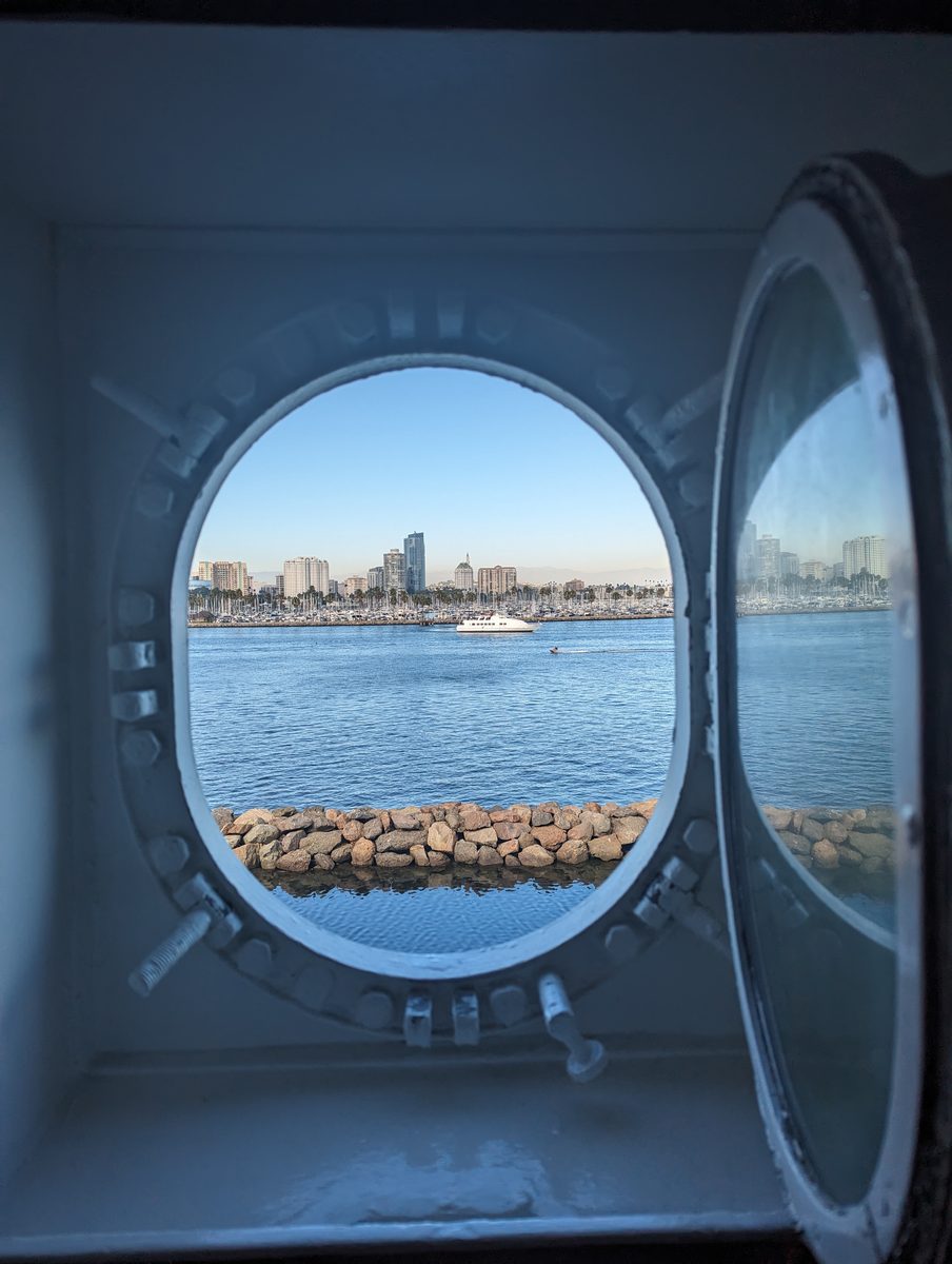 Long Beach skyline and the harbor jetty, framed through a heavy steel porthole of the Queen Mary
