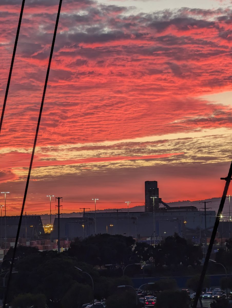 Dramatic red sunset over the Long Beach industrial port, with the Queen Mary's rigging cutting black silhouettes across the sky