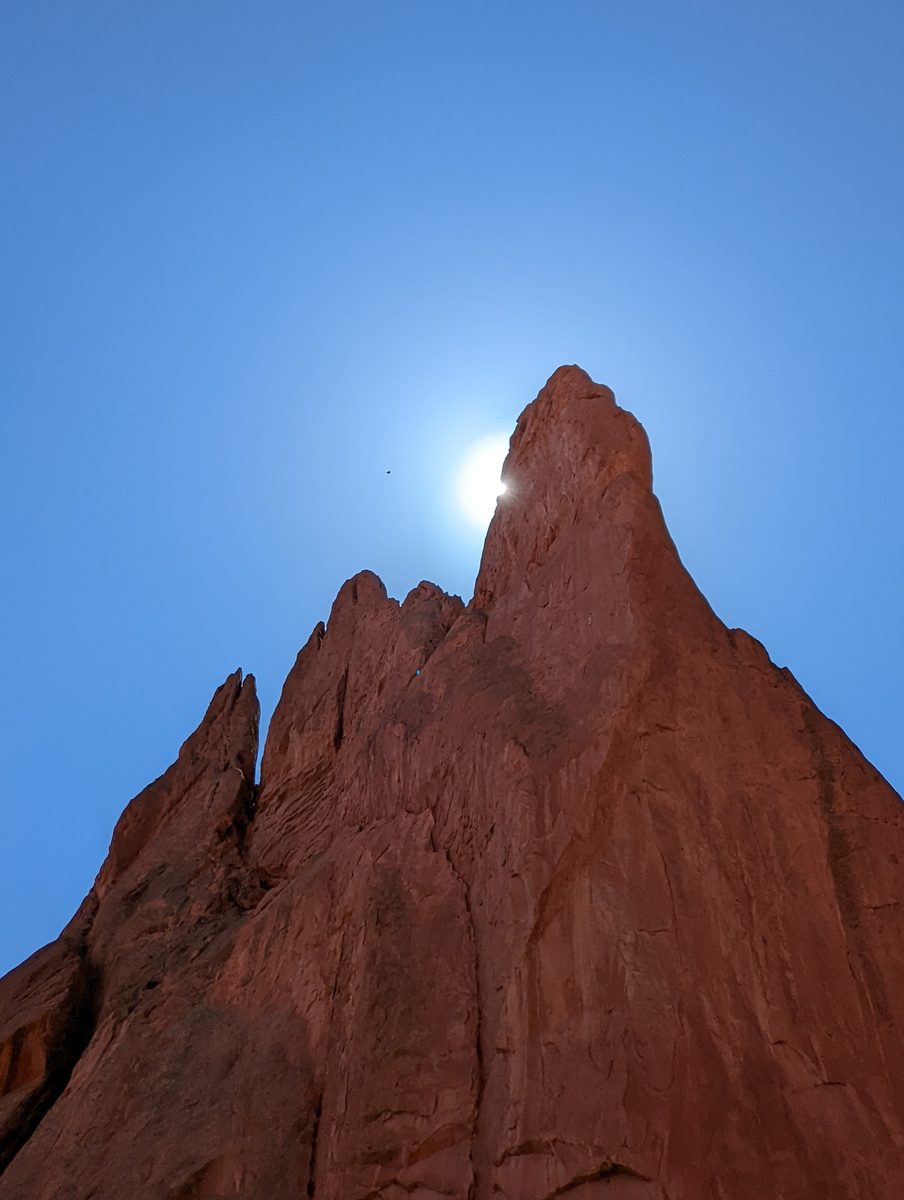 A dramatic backlit view of a jagged red rock spire with the sun peeking out from behind it