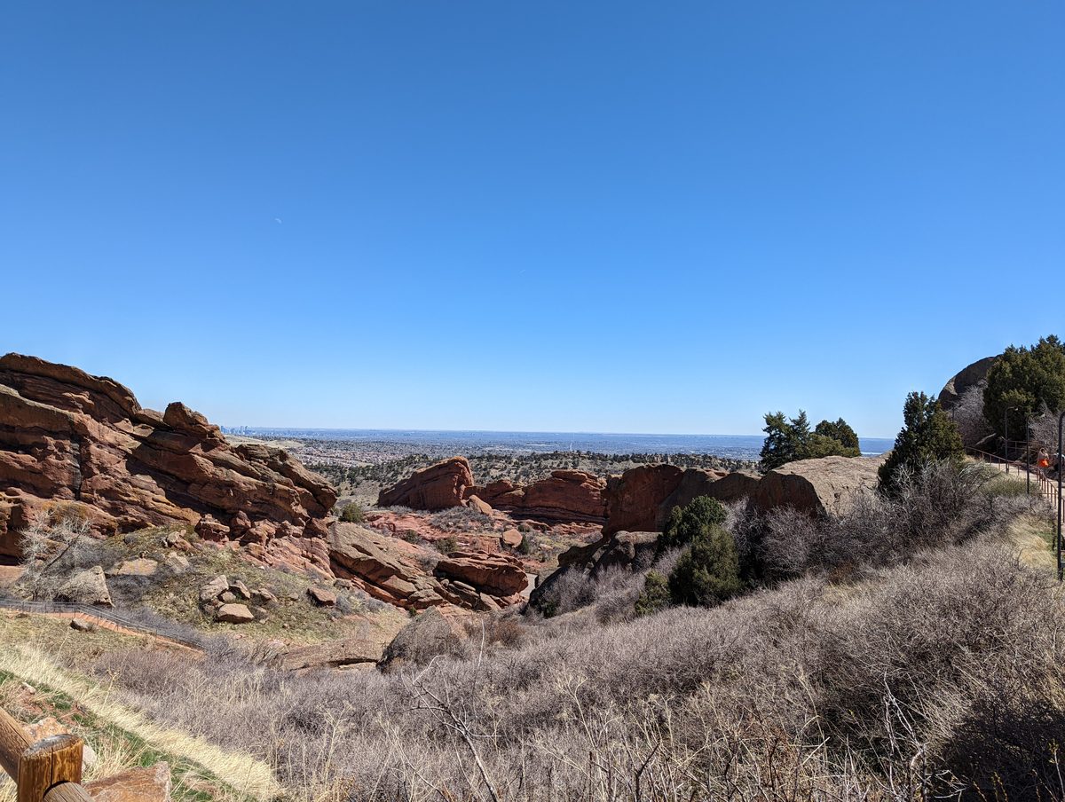A wide overlook of layered red rock formations with the hazy Denver skyline visible on the horizon