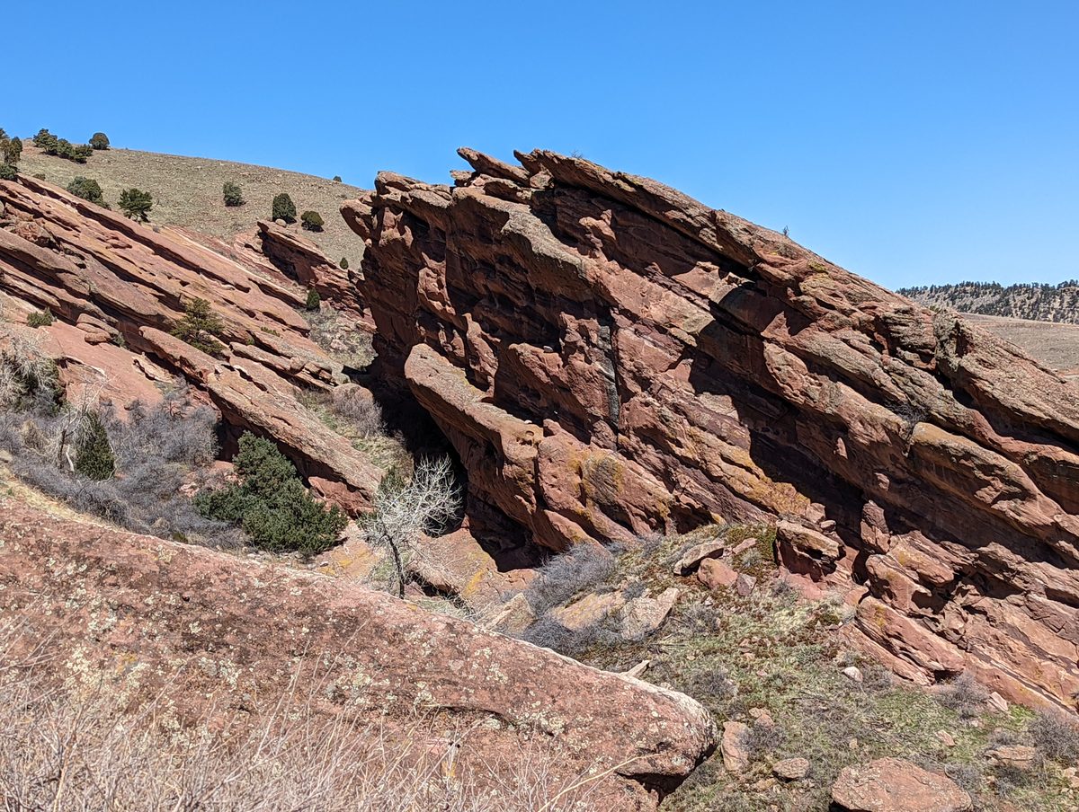A trail-level view of a layered red sandstone formation with sparse junipers