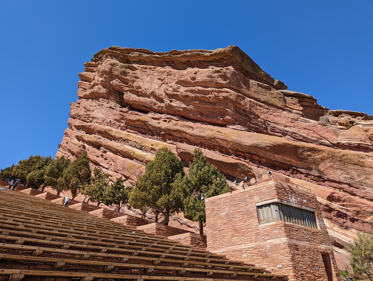 A close view looking up at the rock formation above the amphitheater seating with junipers and a small building