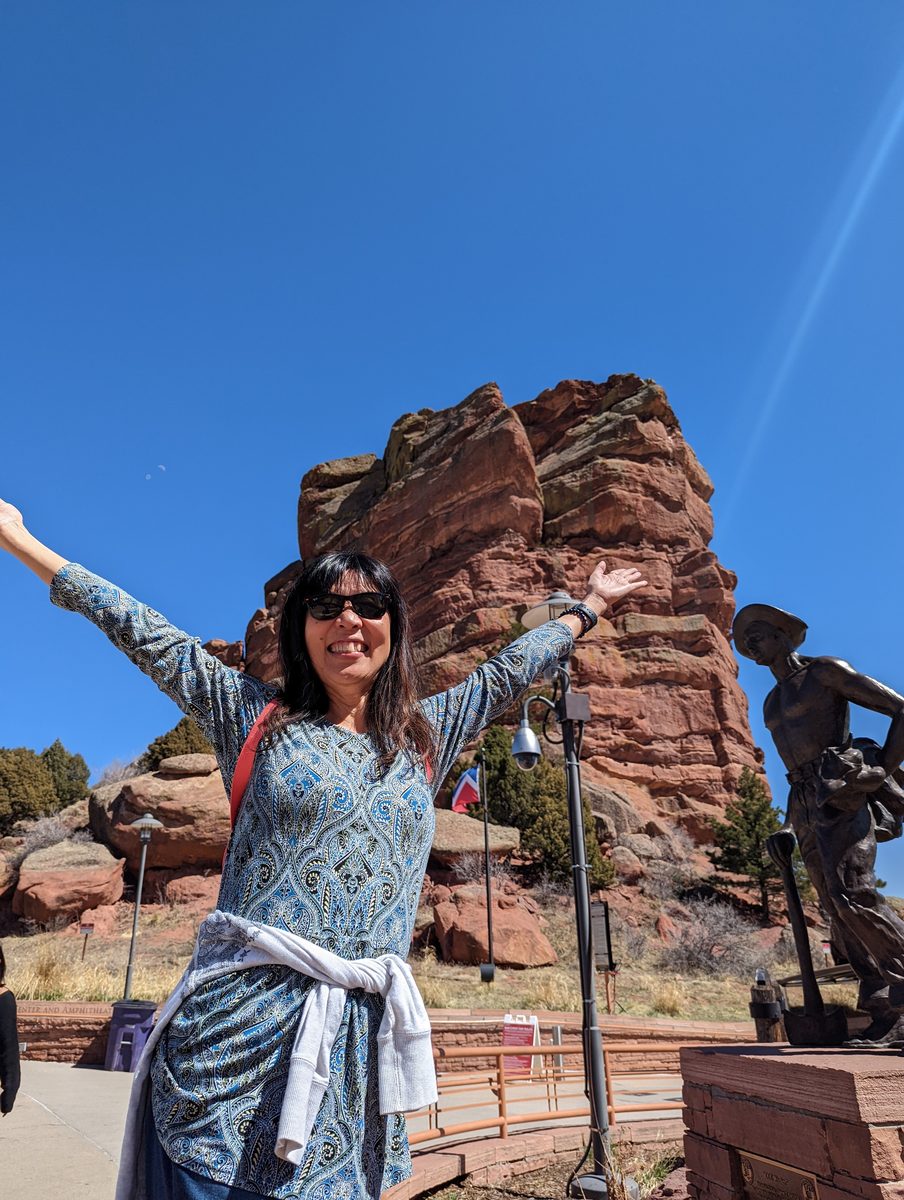 Ako with arms raised triumphantly in front of a red rock spire and a bronze statue near the visitor center entrance