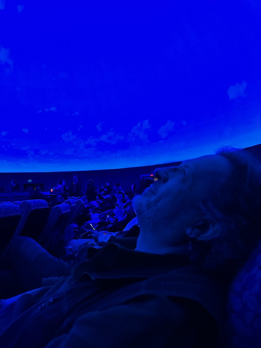 A person reclining and looking up at the blue projected sky inside the domed planetarium theater