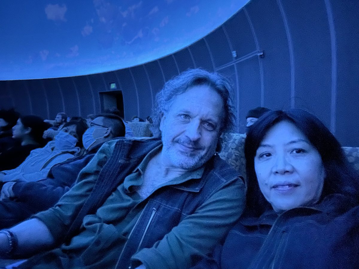 Eddy and Ako selfie inside the blue-lit planetarium dome with other seated visitors around them