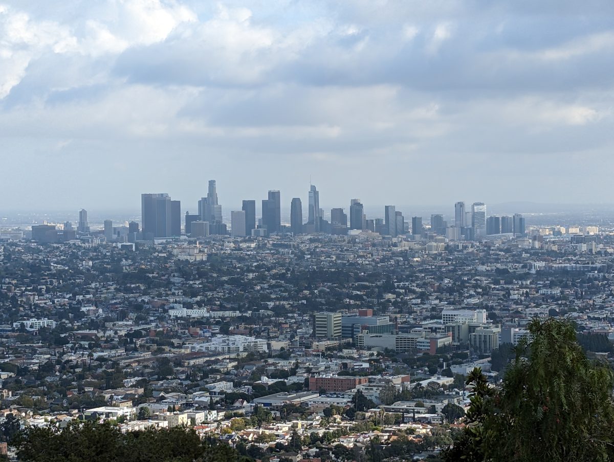 A hazy daytime view of the downtown Los Angeles skyline seen from a hillside above the city