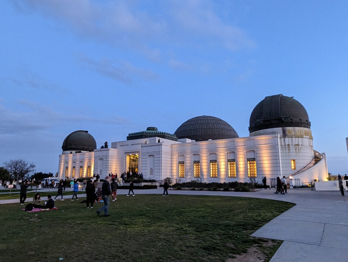 The floodlit Griffith Observatory building with its domes against a darkening blue dusk sky, visitors on the lawn