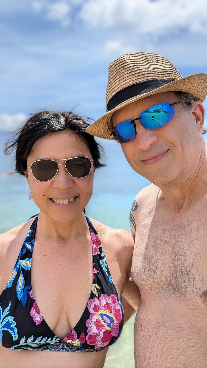Ako and Eddy on Ka'anapali Beach — crystal clear blue-green water behind them, Ako in a colorful bikini, Eddy in a straw hat and blue mirrored sunglasses