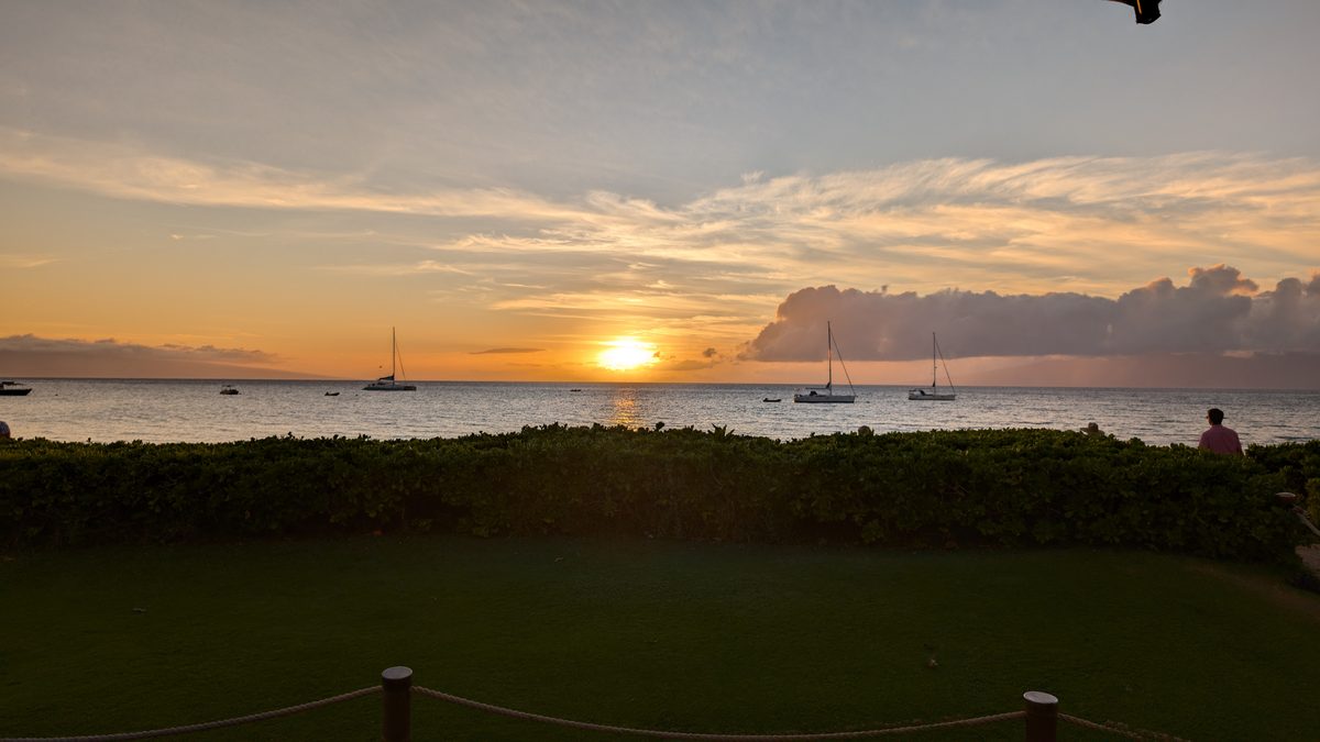 Ka'anapali sunset — sailboats moored on calm water, sun setting orange on the horizon, low hedge in the foreground