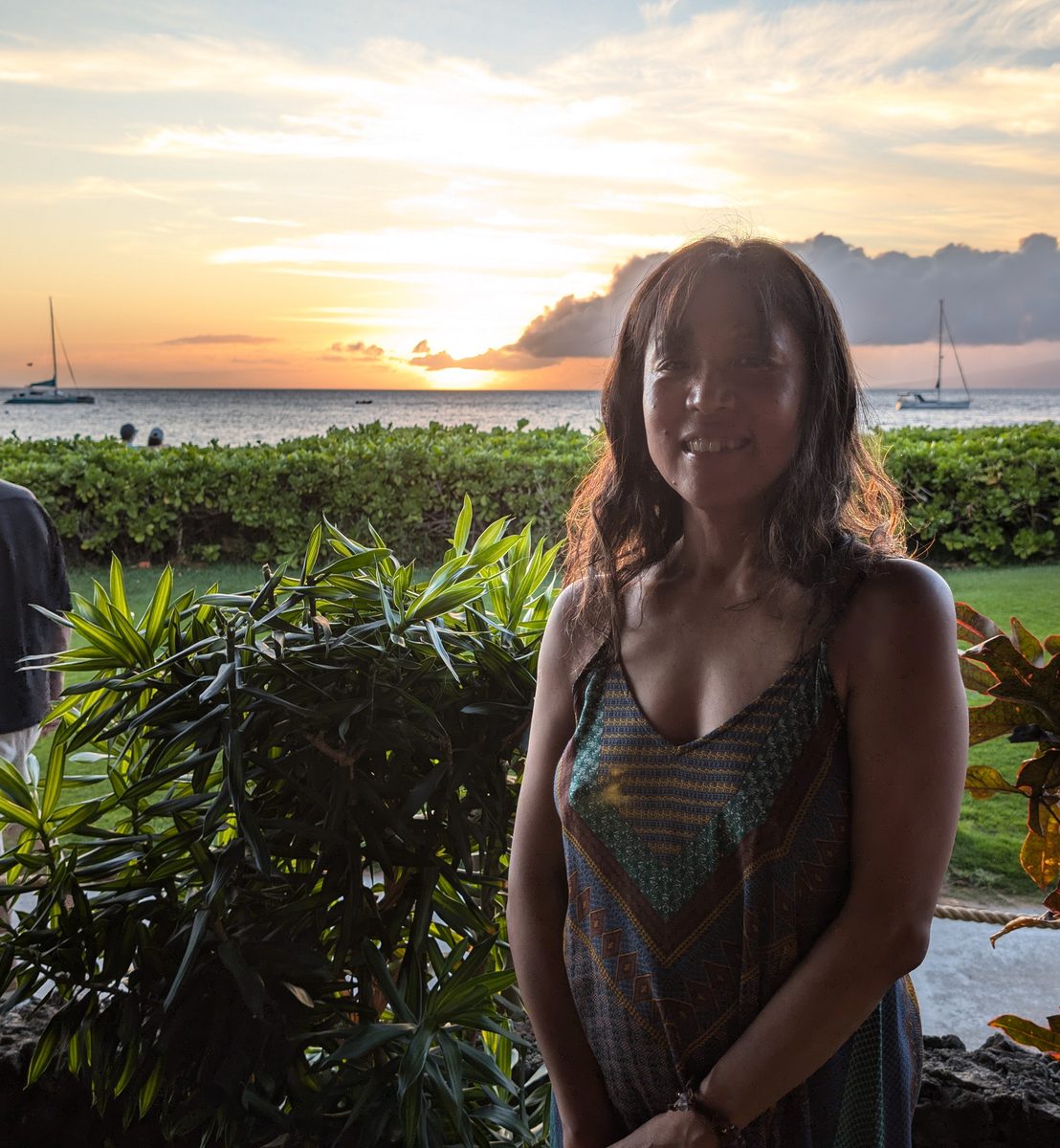 Ako in a colorful patterned dress at the Ka'anapali beachfront, sailboats and sunset behind her