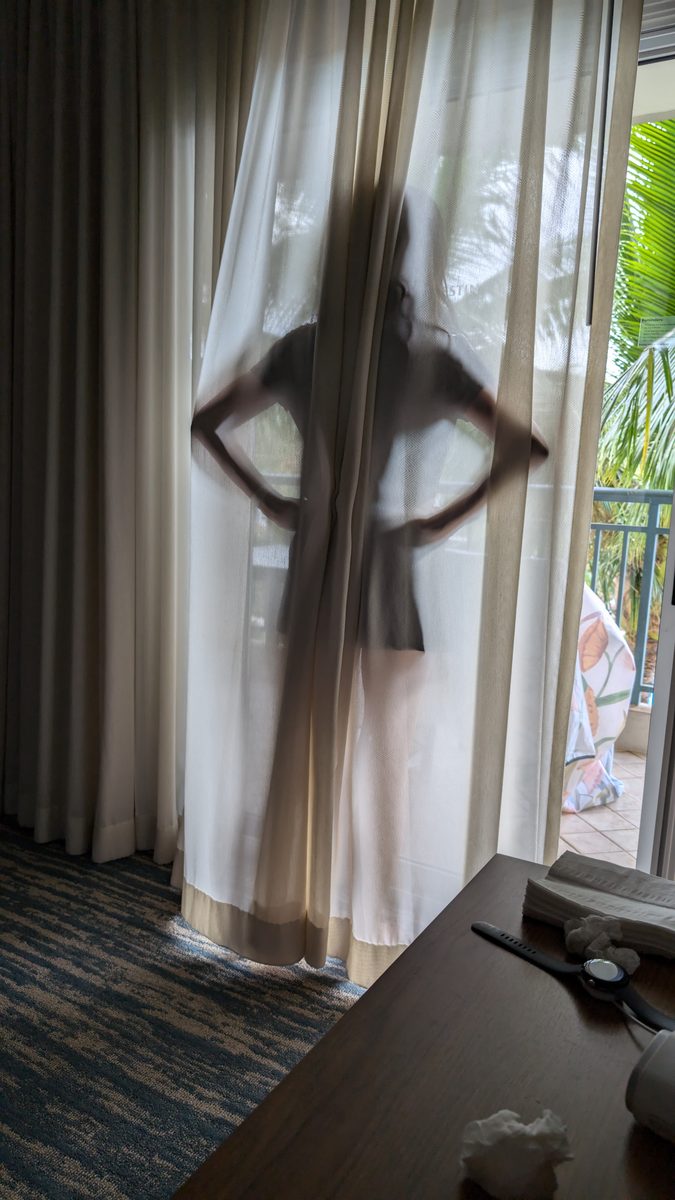 Ako's silhouette seen through sheer white hotel room curtains, palm trees and balcony visible through the glass door behind