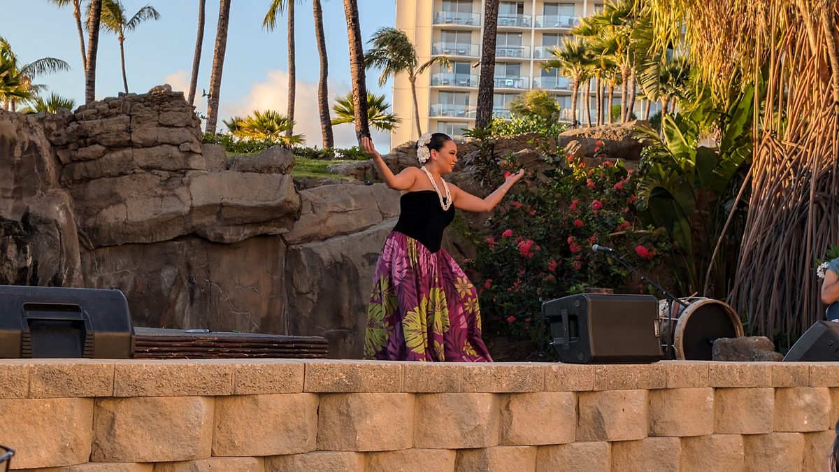 A hula dancer on the luau stage at golden hour — black top and tropical floral skirt, arms extended