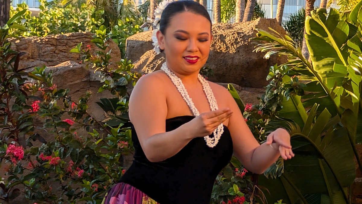 Close portrait of a hula dancer smiling warmly, wearing a white pearl lei necklace, tropical flowers behind her
