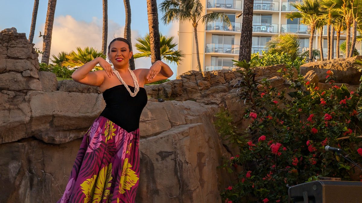 Hula dancer with arms raised and curved behind her, colorful tropical skirt, palm trees behind