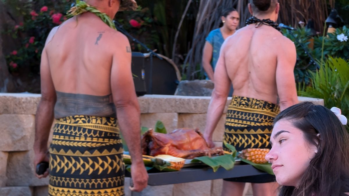 Two men in traditional Polynesian tapa cloth malo carrying a whole roasted pig on banana leaves and pineapples