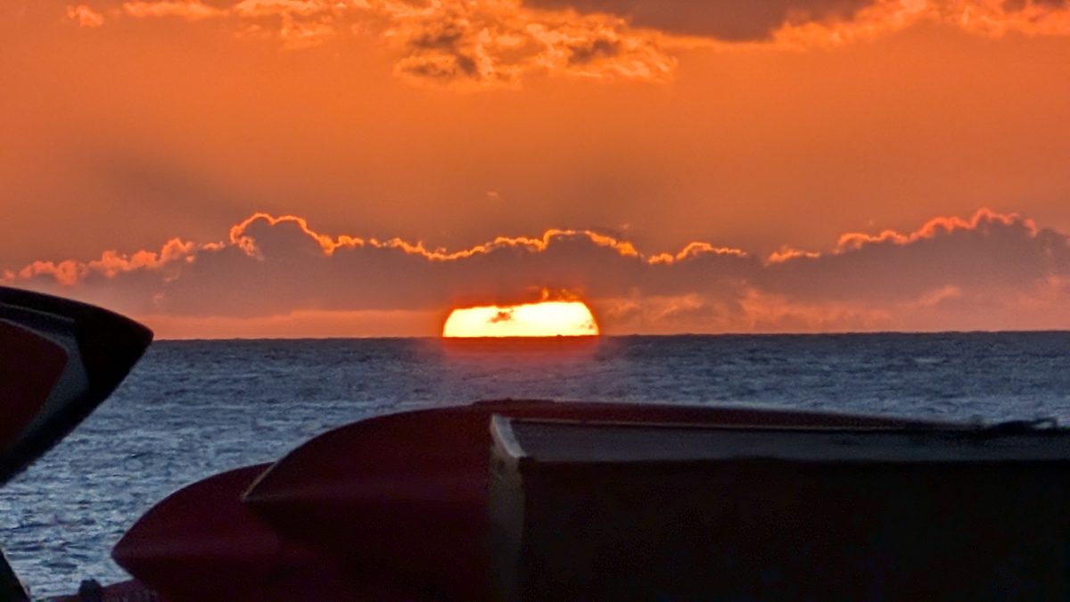 Brilliant orange luau sunset — the sun piercing a gap in the clouds, a boat silhouette in the foreground