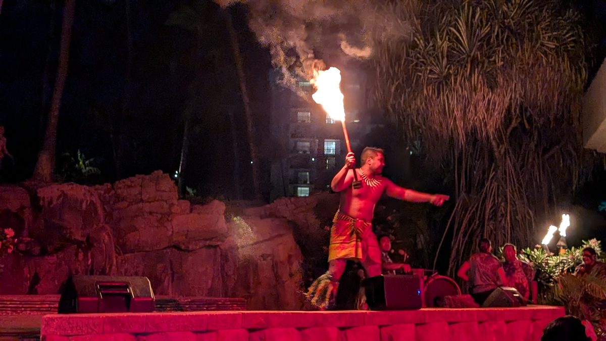 Fire knife dancer holding a blazing torch overhead on the luau stage, red lighting all around