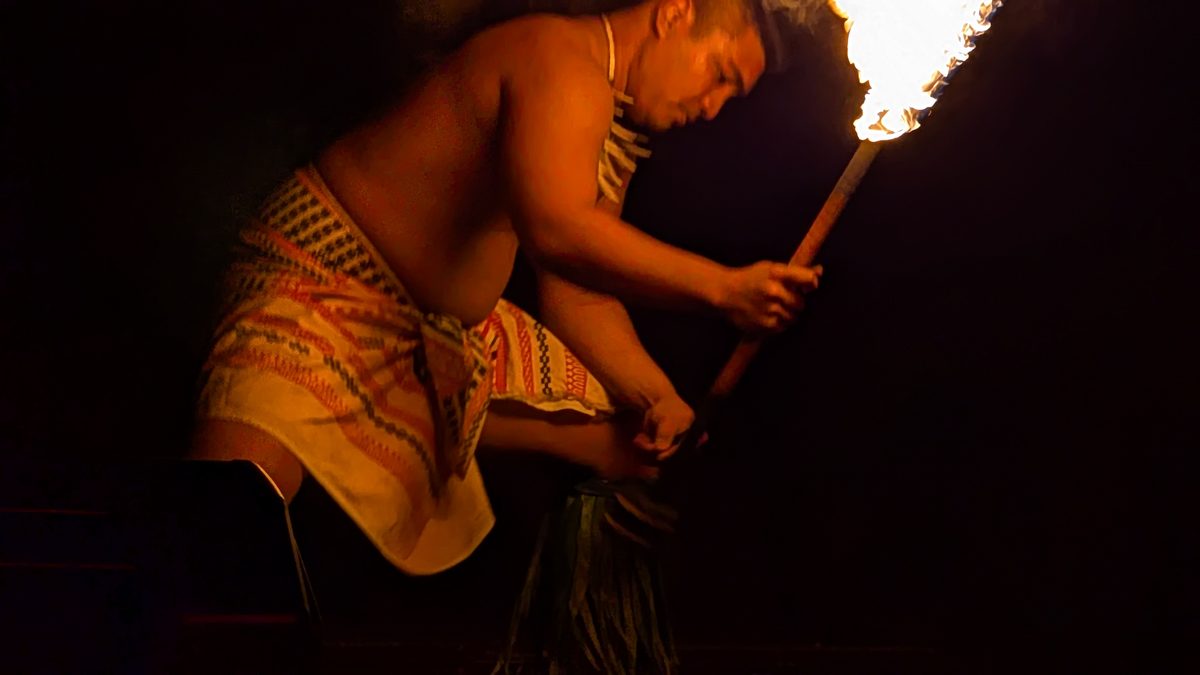 Fire knife dancer bent low, face illuminated by the flame, tapa cloth skirt visible in the darkness