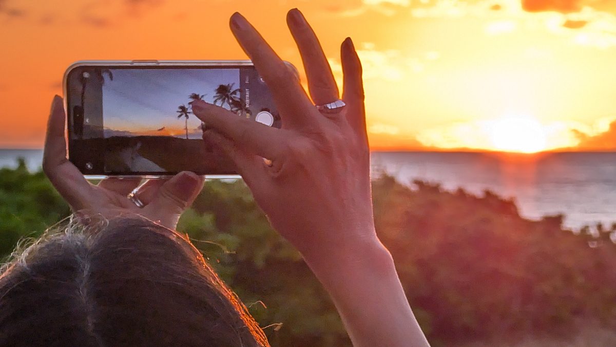 Ako's hands holding her phone up to photograph a palm tree sunset — wedding ring clearly visible