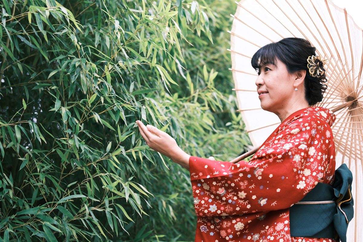 Ako in her red furisode, reaching toward bamboo leaves, the wagasa open beside her — a serene profile