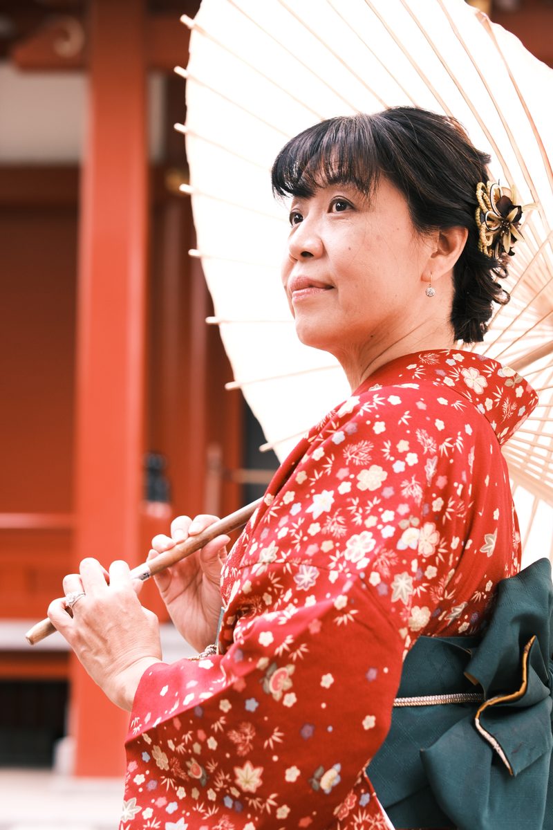 Close-up of Ako's face tilted back, wagasa handle near her cheek, vermilion temple wall behind her