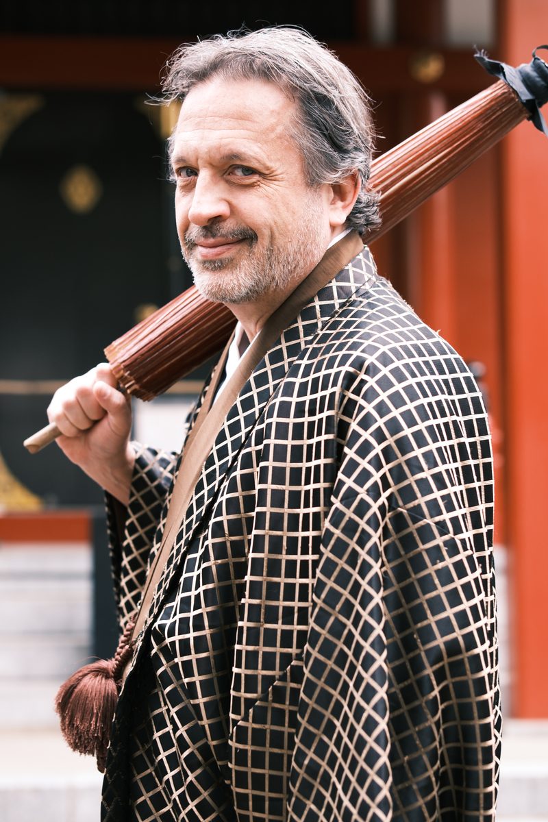 Close-up portrait of Eddy smiling, holding a folding sensu fan, vermilion temple detail behind him