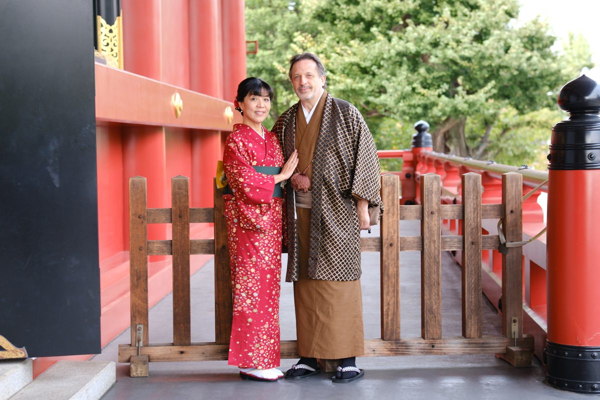 Eddy and Ako side by side on the Sensoji vermilion bridge railing, warm smiles, autumn trees behind