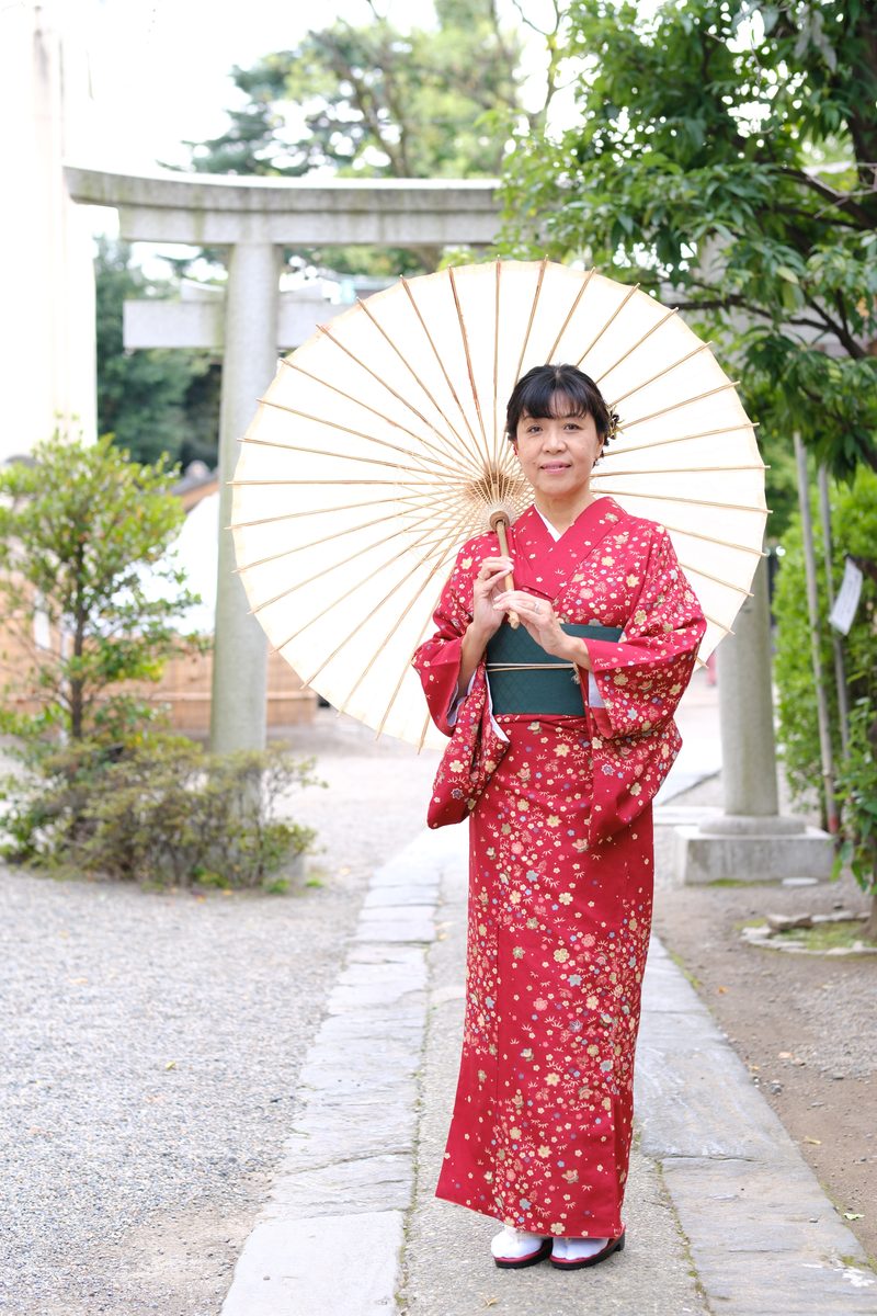 Ako full-length in her red furisode by a stone wall, wagasa in hand, a small red torii gate behind her