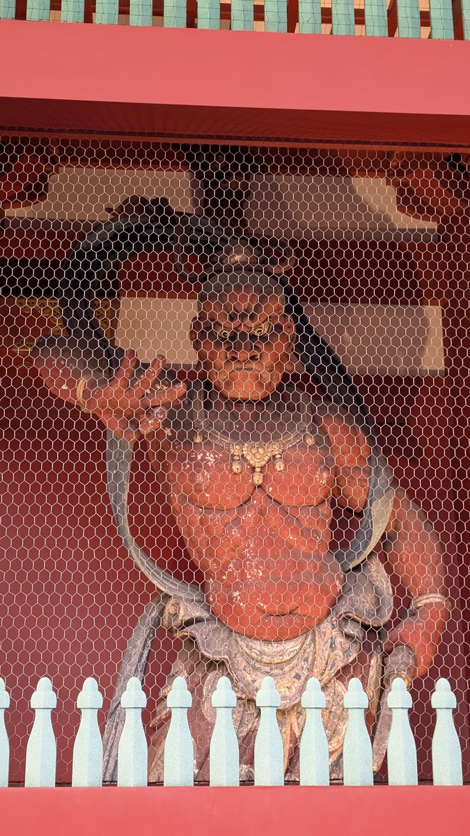 A fierce vermilion guardian deity statue behind the wire mesh of the Hozomon gate