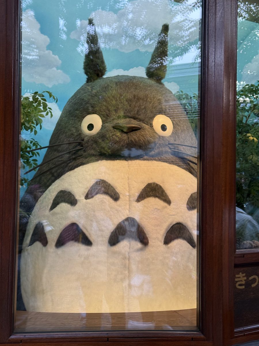 A giant plush Totoro figure seen through a museum window, whiskers and all
