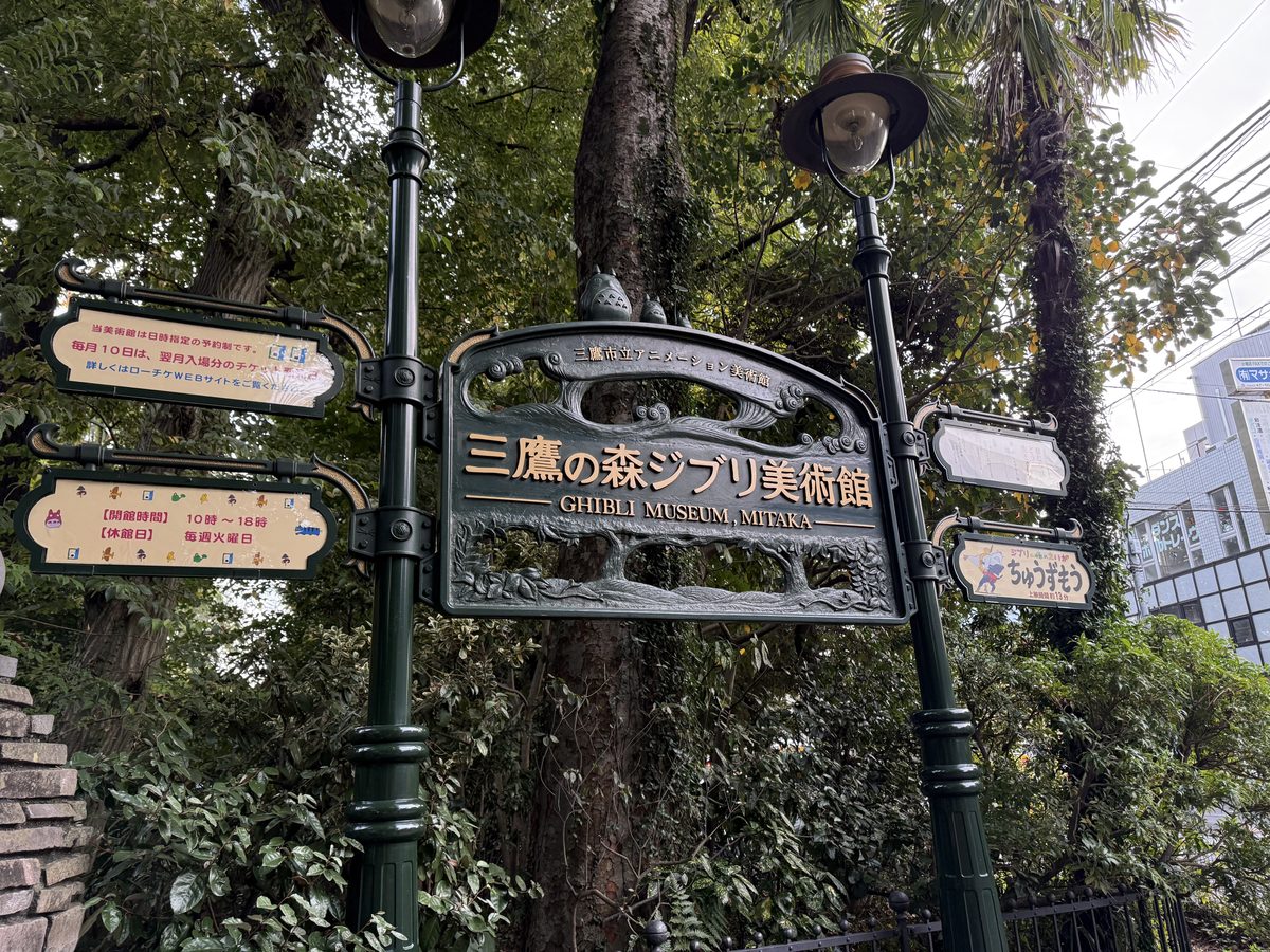 An ornate cast-iron signpost reading Ghibli Museum, Mitaka with small Totoro figures on top
