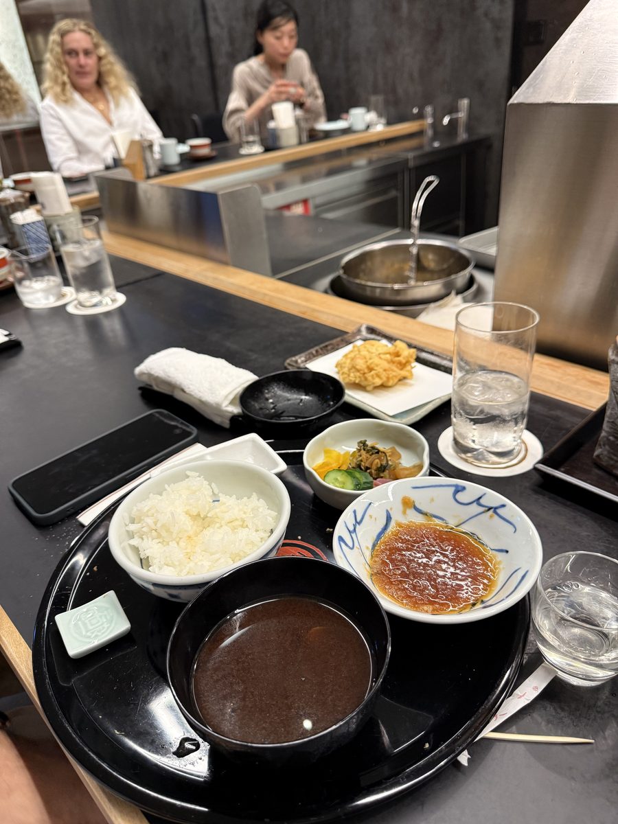 A tempura counter meal with rice, miso soup, pickles and kakiage, other diners visible across the bar