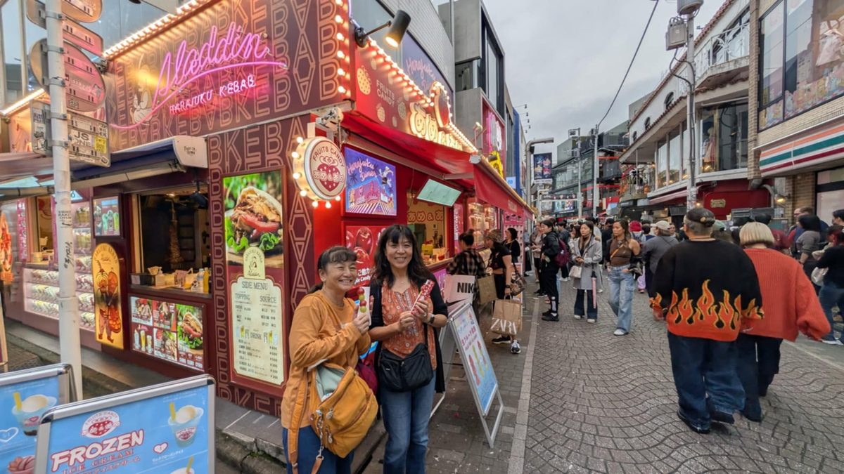 Two friends smiling and holding candied fruit skewers outside a Harajuku kebab and crepe stand
