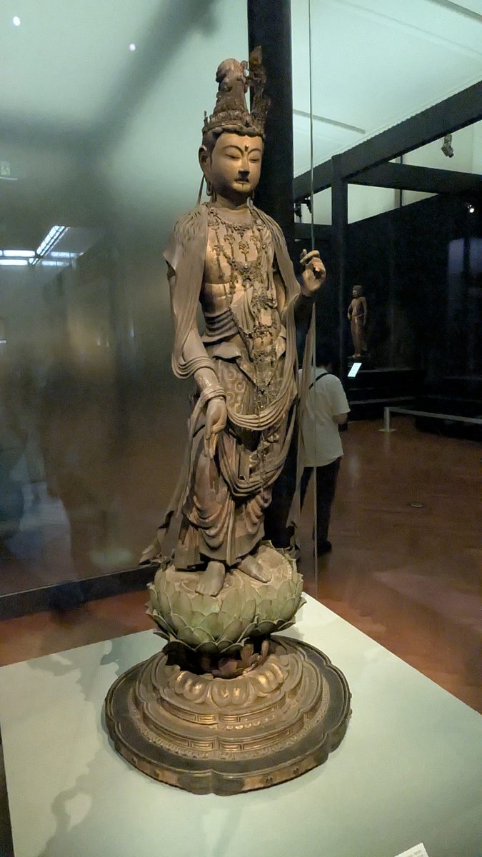 A standing wooden bodhisattva statue on a lotus pedestal in a museum gallery