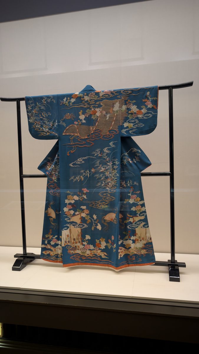 A blue silk kimono with floral designs spread on a T-stand in a glass case