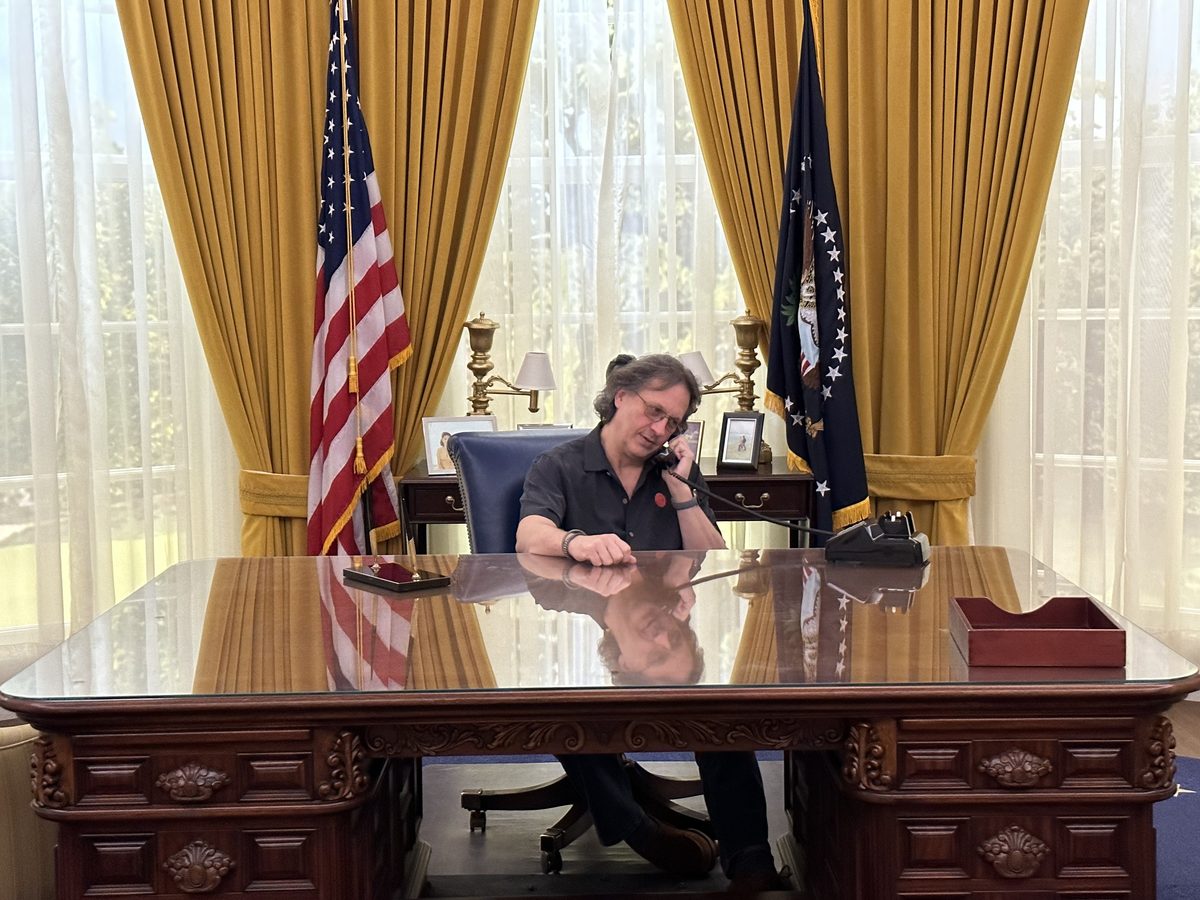Eddy seated at the Oval Office desk holding a phone, American flag and curtains behind him