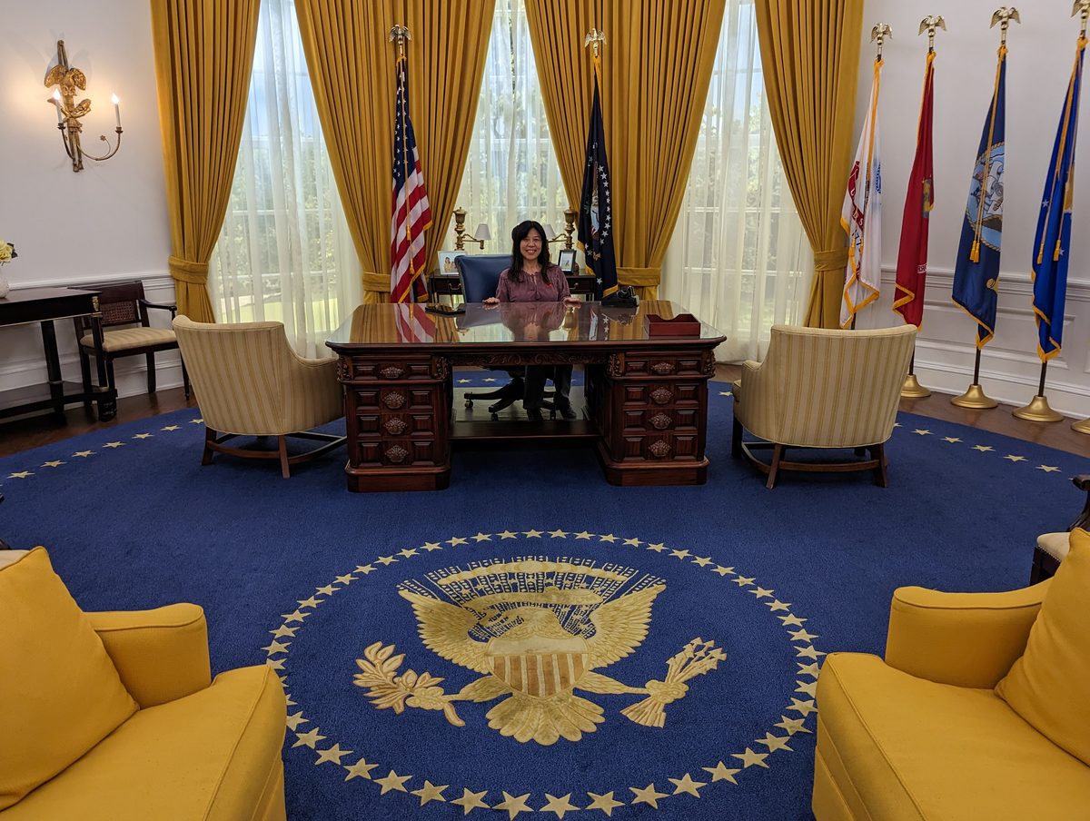 Ako seated at the Resolute desk in the full-size Oval Office replica, gold drapes and presidential rug