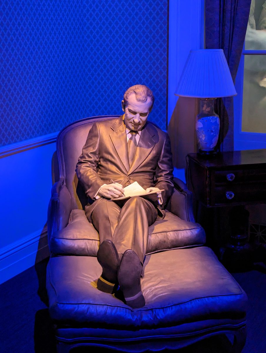 A lifelike statue of Nixon seated in an armchair writing on a notepad under blue lamplight